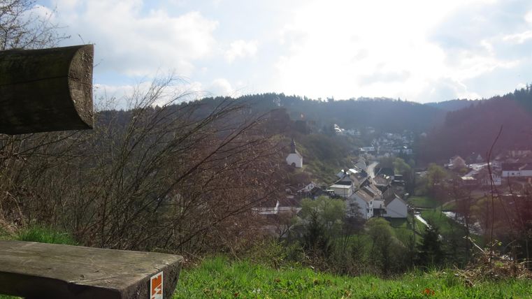 A picturesque view of a small village in a valley, surrounded by wooded hills. The sky is partly cloudy and the sun is shining through the clouds.