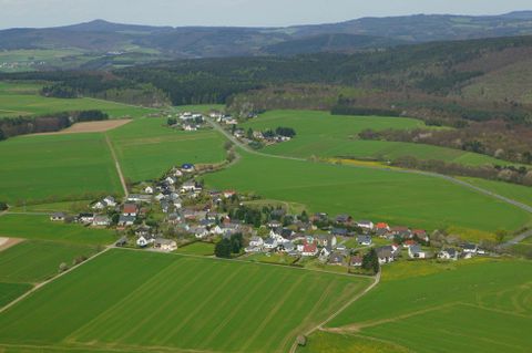 A picturesque rural landscape with a small village surrounded by lush greenery. In the background, gentle hills and a clear sky can be seen.