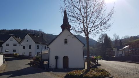 Eine kleine, weiße Kapelle mit einem spitzen Turm steht am Straßenrand. Im Hintergrund sind die Berge und einige Wohnhäuser zu sehen.