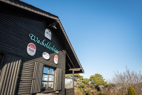 A black wooden hut with the inscription "Wacholderhütte" and several signs on the wall. The sky is clear and blue, surrounded by gentle trees.