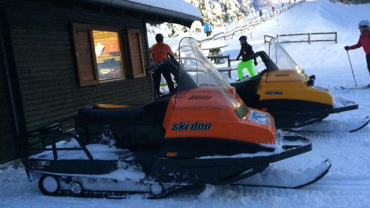 Zwei Schneemobile stehen neben einem Holzhaus in einer verschneiten Landschaft. Im Hintergrund sind Skifahrer und schneebedeckte Bäume zu sehen.