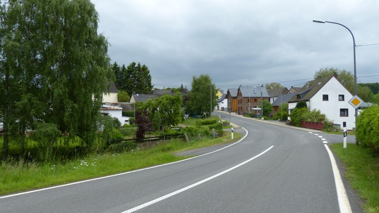 A winding road leads through a quiet rural setting. In the background, some houses and plenty of greenery can be seen.