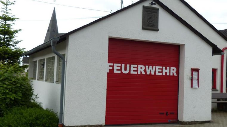 Ein Feuerwehrgebäude mit einem roten Garagentor. Das Haus hat eine weiße Fassade und ist von Pflanzen umgeben.