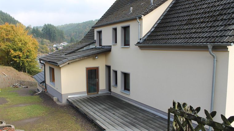 Ein modernes Haus mit einer Holzterrasse und einem Garten. Im Hintergrund sind bewaldete Hügel zu sehen.