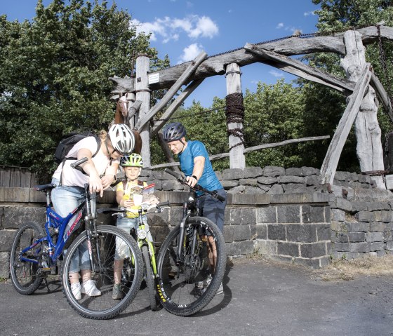 Une famille &agrave; v&eacute;lo regarde une carte devant un &eacute;quipement minier historique en bois. Le ciel est bleu et l'environnement vert., &copy; Klaus-Peter Kappest/Vulkanregion Laacher See