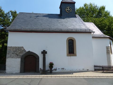 Eine kleine, weiße Kirche mit einem grauen Dach und einer Uhr an der Front. Neben der Kirche steht ein Kreuz und Blumen sind am Eingang platziert.