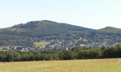 A green landscape with gentle hills and a small village in the foreground. The sky is clear and sunny.