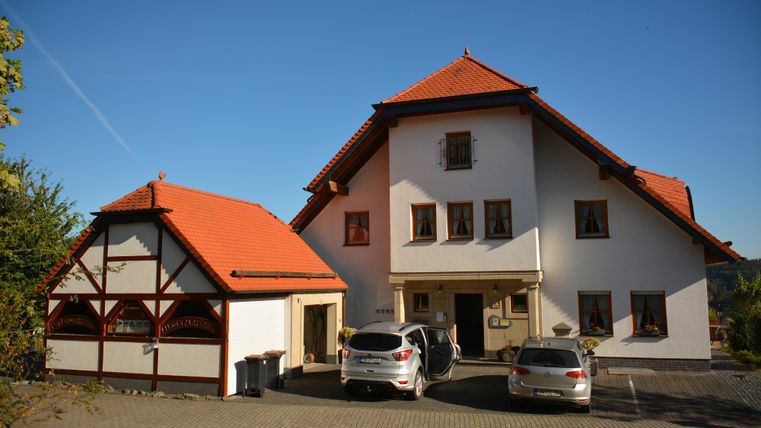 Ein schönes, modernes Haus mit rotem Dach und einem Nebengebäude. Der Parkplatz ist mit mehreren Autos belegt und der Himmel ist klar und blau.