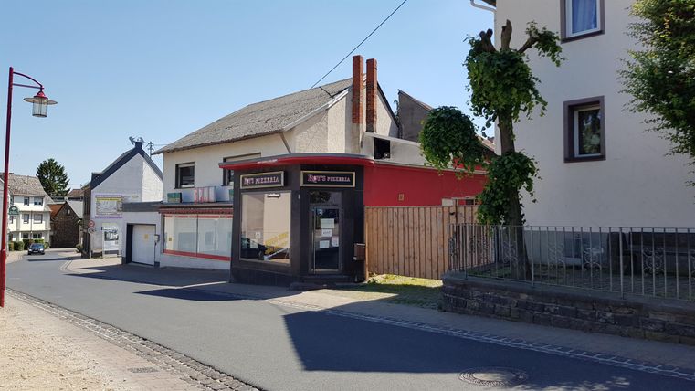 Eine ruhige Straße mit einem kleinen, roten Gebäude und benachbarten Wohnhäusern. Der Himmel ist klar und blau.