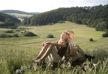 Zwei Personen entspannen auf einer Bank vor einer gr&uuml;nen, h&uuml;geligen Landschaft mit W&auml;ldern im Hintergrund., &copy; Traumpfade
