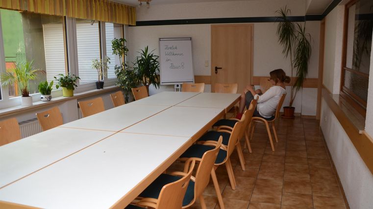Ein leerer Besprechungsraum mit einem langen Tisch und Holzstühlen. Eine Person sitzt in einer Ecke und schaut auf ihr Handy.