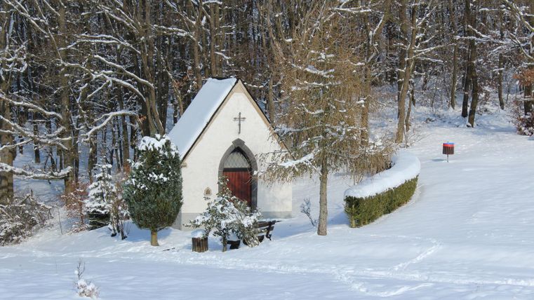 Eine kleine Kapelle im Schnee, umgeben von Bäumen. Die Landschaft ist friedlich und winterlich.