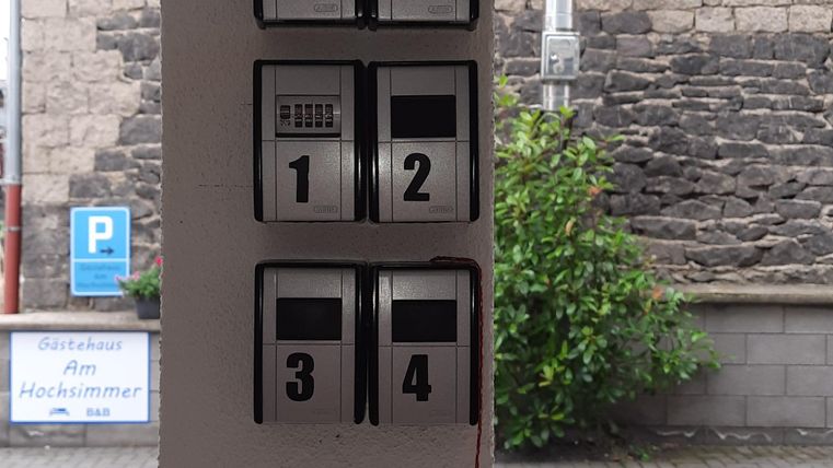 A black information system with several switches is mounted on a wall. In the background, green plants and a stone wall can be seen.