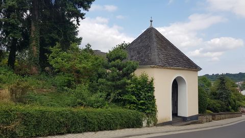 Eine kleine Kapelle mit einem runden Dach steht umgeben von Bäumen und Sträuchern. Der Himmel ist teilweise bewölkt und die Umgebung ist grün.