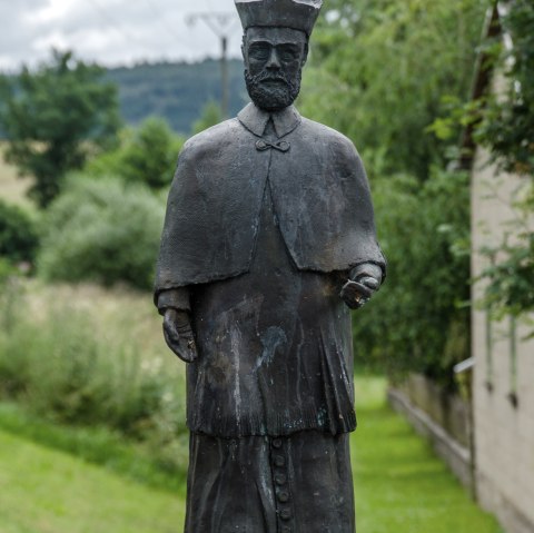 Bronzen beeld van een man in traditionele kleding op een sokkel, omringd door een groen landschap en een bewolkte lucht., &copy; Volker Windheuser