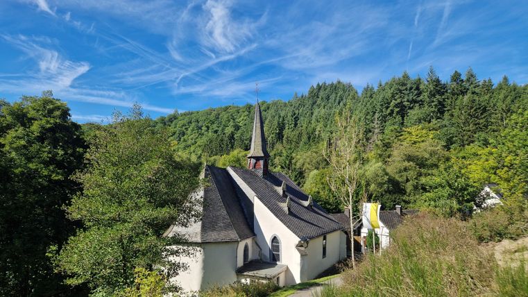 Eine malerische Kirche umgeben von Bäumen und Hügeln. Der Himmel ist klar mit einigen Wolken.