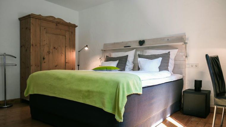 A cozy bedroom with a large bed and a green blanket. In the room, there is a wooden wardrobe and a modern chair.