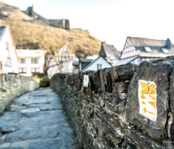 Wanderweg-Markierung auf einer Steinmauer, im Hintergrund Fachwerkh&auml;user und eine Burg auf einem H&uuml;gel., &copy; Eifel Tourismus GmbH, D. Ketz