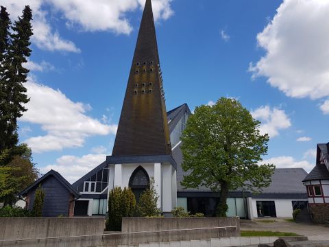 Eine moderne Kirche mit einem hohen, spitzen Turm und großen Fenstern. Im Vordergrund steht ein grüner Baum und die Umgebung ist von leicht bewölktem Himmel umgeben.