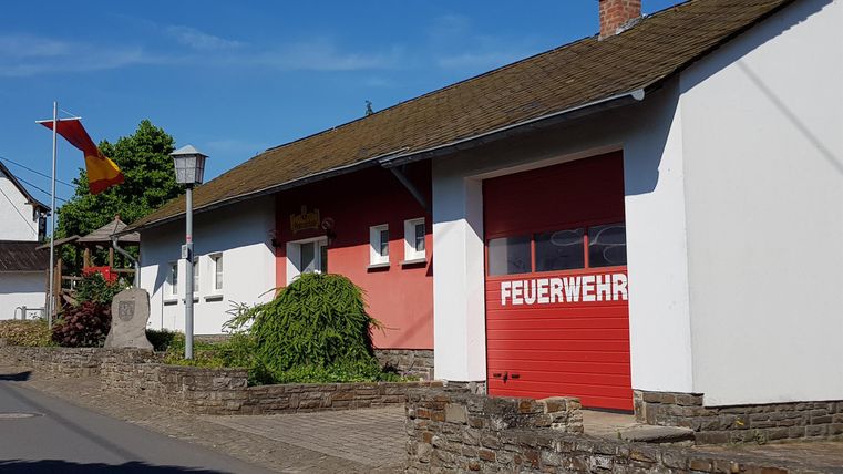 Eine Feuerwehrstation mit rotem Tor und hellen Wänden. Vor der Station sind einige Bäume und eine Fahne sichtbar.