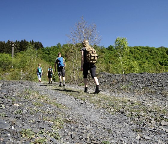 Eine Gruppe von Wanderern mit Rucks&auml;cken geht einen Schieferweg entlang, umgeben von gr&uuml;ner Vegetation und blauem Himmel., &copy; Laura Rinneburger