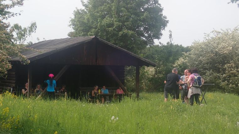 A small group of people is gathered in front of a wooden shelter in a green meadow. In the background, trees and flowering shrubs are visible.