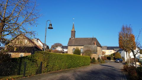 Eine malerische Straße mit einer Kirche im Hintergrund und klar blauem Himmel. Rund um die Straße sind gepflegte Hecken und Häuser zu sehen.