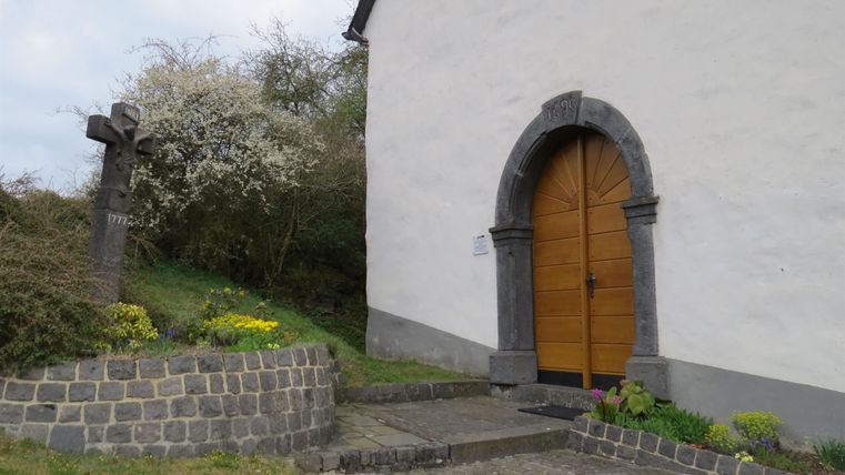 Eine Kirche mit einer schönen Eingangstür aus Holz und einem kleinen Kreuz im Garten. Umgeben von Blumen und grüner Vegetation.