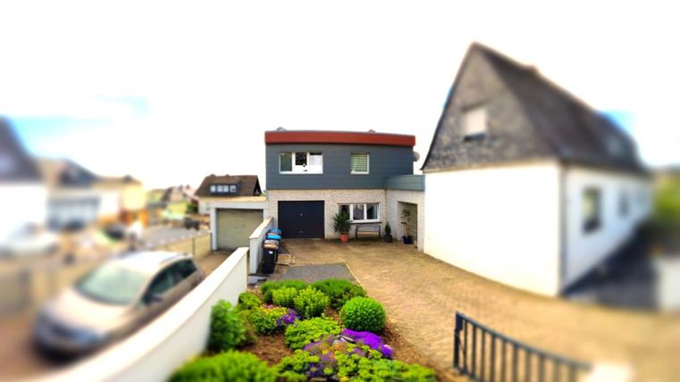 Ein modernes Haus mit einem Garten voller bunter Blumen. Die Auffahrt ist gepflastert und es stehen mehrere Fahrzeuge in der Nähe.
