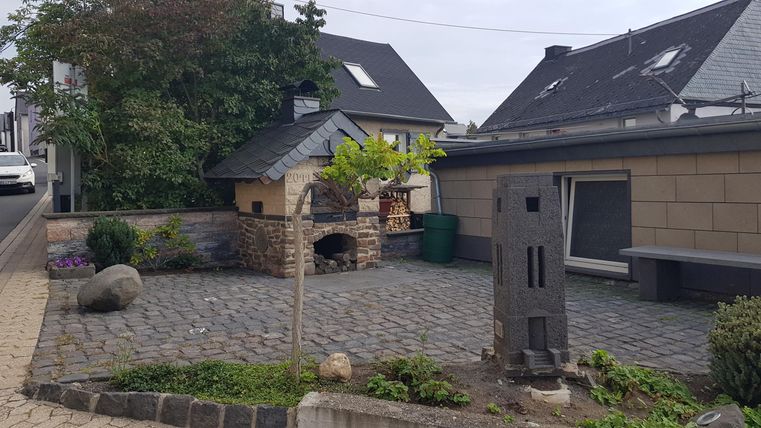 Ein gepflegter Vorgarten mit gepflastertem Weg und grünen Pflanzen. Im Hintergrund sind moderne Häuser und ein kleiner Grillplatz zu sehen.