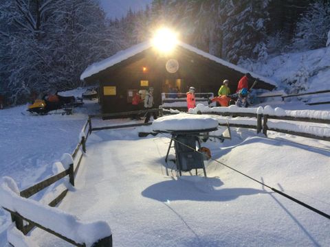 Eine gemütliche Berghütte im Schnee mit einem hellen Licht über dem Eingang. Die Umgebung ist von frischem, weißem Schnee und Bäumen umgeben.