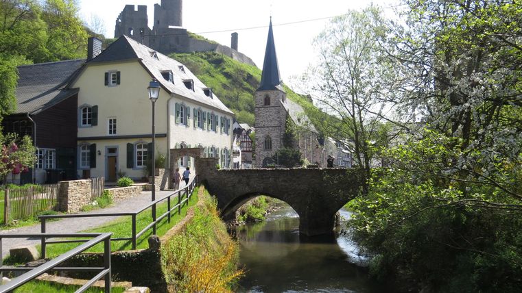 Eine malerische Landschaft mit einem kleinen Fluss und einer alten Steinbrücke. Im Hintergrund sind Häuser und eine Burg auf einem Hügel zu sehen.