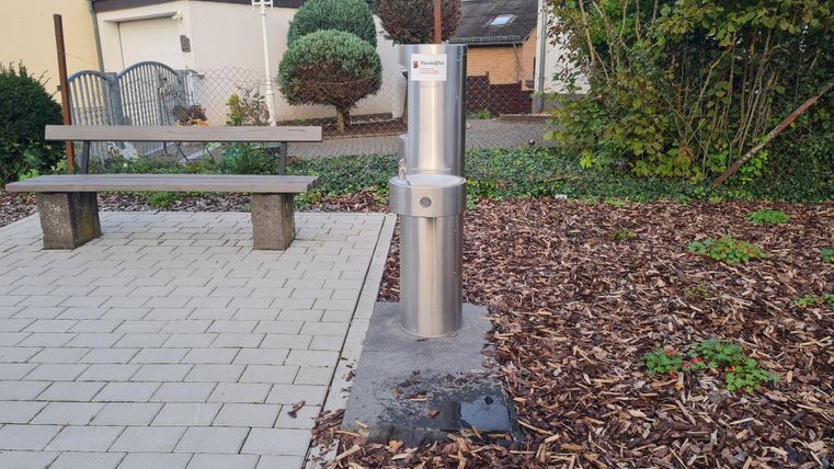 A stainless steel water fountain stands in a small park. Nearby, there is a bench and well-tended flower beds.
