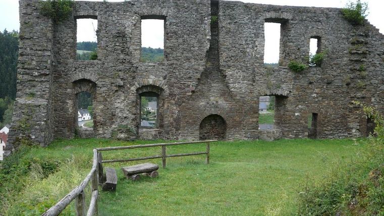 Eine alte Ruine aus Stein, umgeben von grünem Gras. Im Hintergrund sind Hügel und eine kleine Stadt zu sehen.