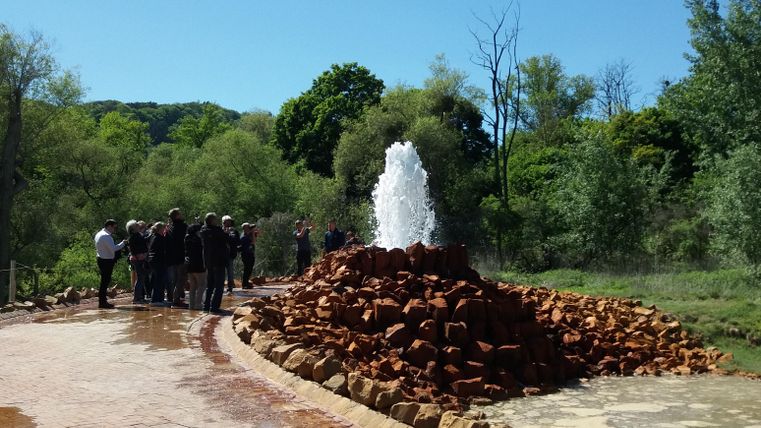 A bubbling fountain surrounded by stones and trees. A group of people stands nearby and enjoys the view.