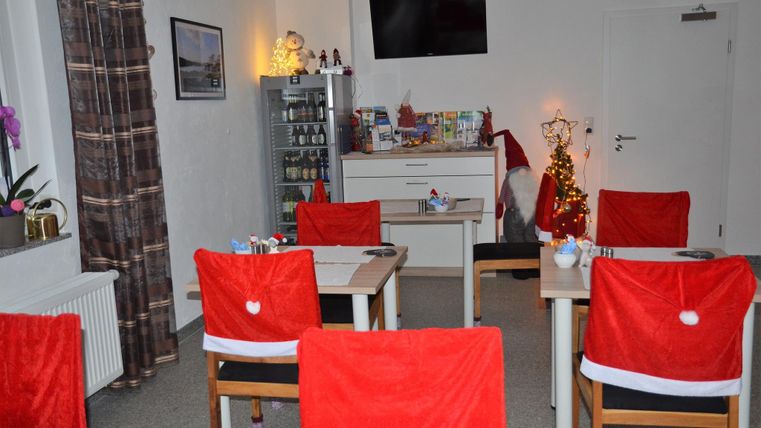 A cozy room with tables and chairs decorated with red Christmas coverings. In the background stands a small Christmas tree and festive decorations are visible.