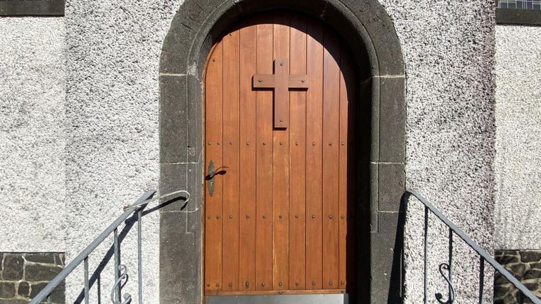Eine Holztür mit einem Kreuz in einem geschwungenen Torbogen. Die Tür befindet sich an einem Gebäude mit einer groben Steinstruktur.