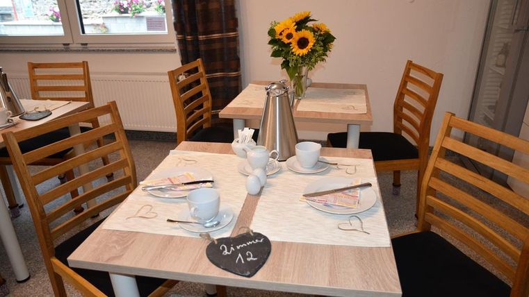 A cozy table with wooden chairs in a bright room. On the table are a coffee pot, cups, and a bouquet of sunflowers.