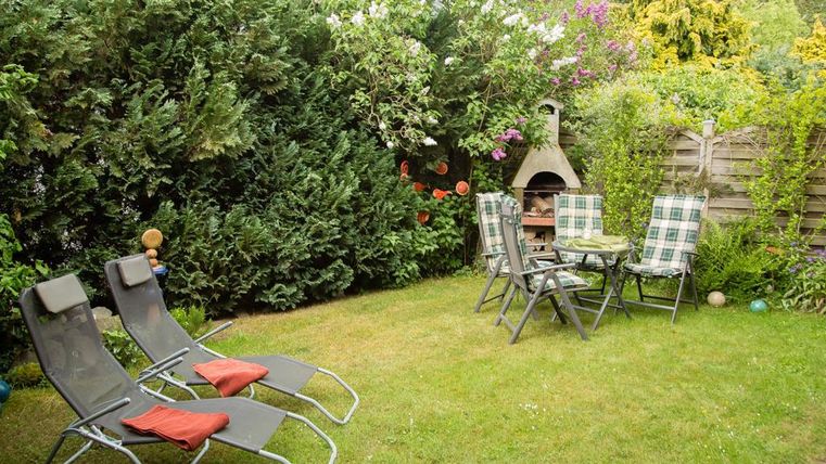 Ein gemütlicher Garten mit zwei Sonnenliegen und einem Tisch mit Stühlen. Im Hintergrund sind hohe Pflanzen und ein Grill zu sehen.