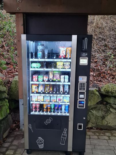 Ein Verkaufsautomat steht unter einem Überstand. Er bietet eine Auswahl an Snacks und Getränken.