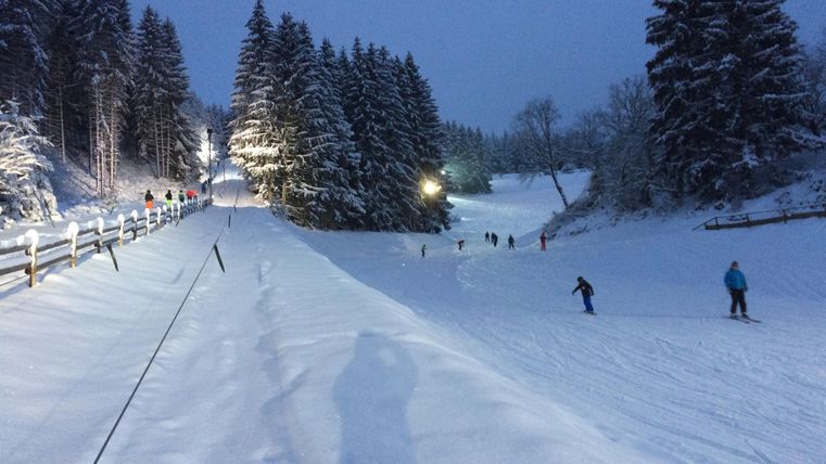 Eine winterliche Landschaft mit viel Schnee und beleuchteten Bäumen. Menschen skifahren und genießen die Freizeit im Freien.