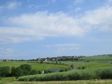 Eine malerische Landschaft mit sanften Hügeln und einem kleinen Dorf im Hintergrund. Der Himmel ist blau und leicht bewölkt.